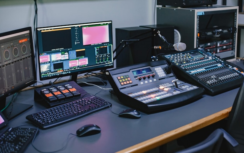 A broadcasting studio setup featuring two computer monitors displaying colorful graphics, a mixing console with various buttons and sliders, and a keyboard and mouse on the desk.
